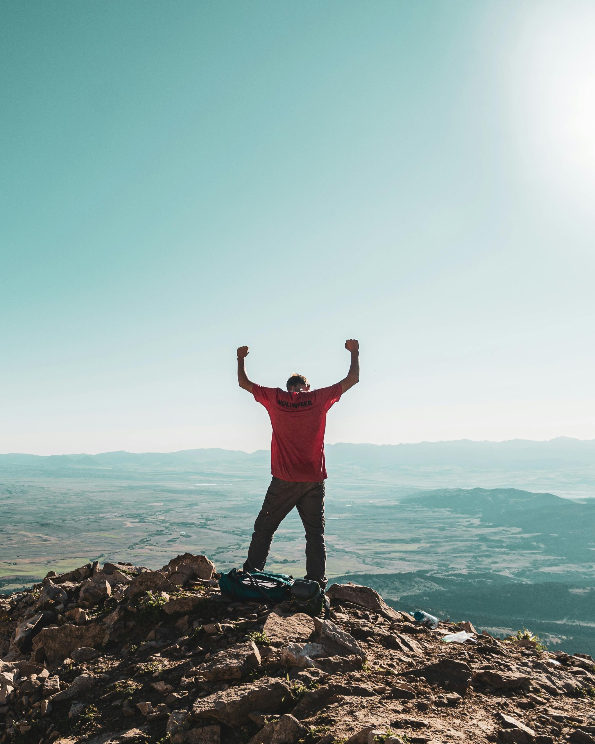 A man triumphantly celebrates on a rocky mountain summit, overlooking vast landscapes under clear skies.